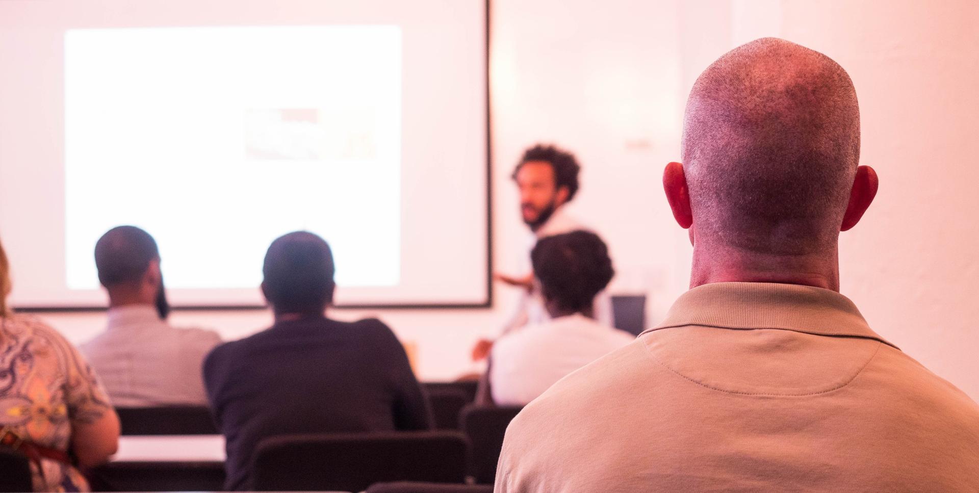Brandon Campbell-Kearns, seen from the rear of a room, teaching digital skills to a group of professionals, with a focus on web development.